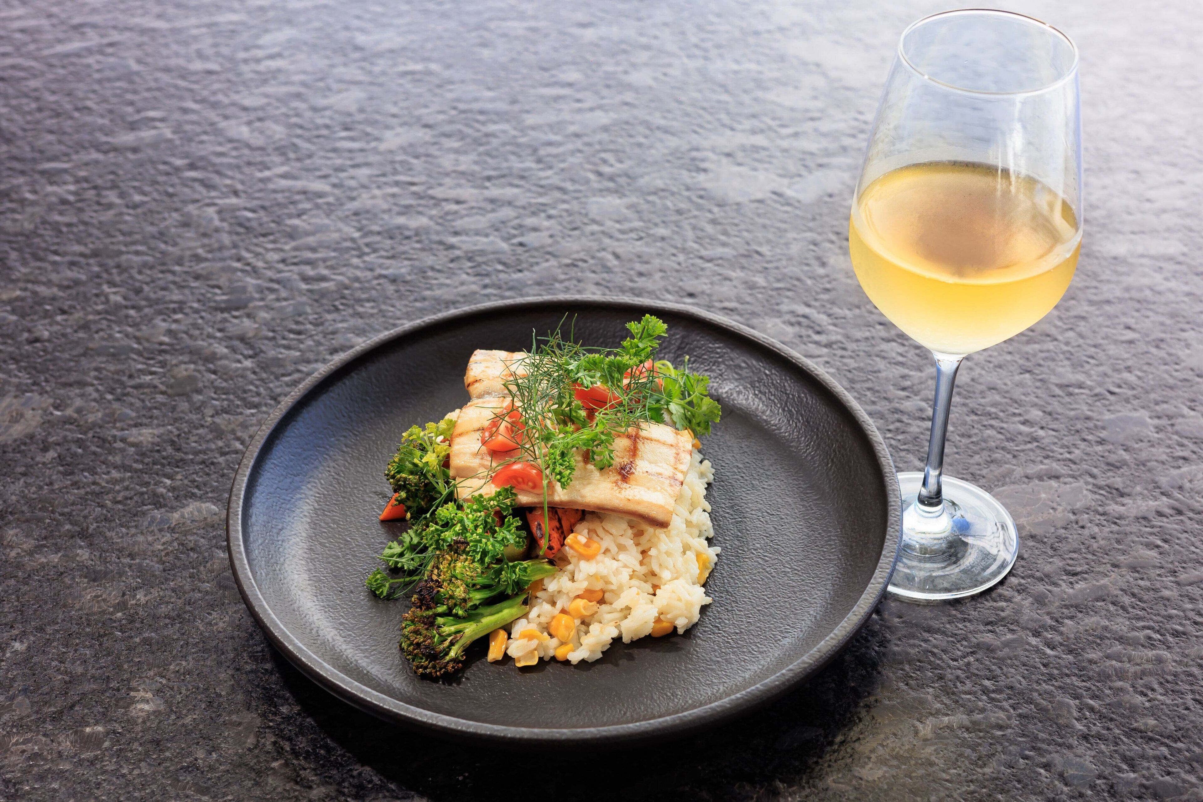 A plate of food with rice, vegetables, and a piece of meat, accompanied by a glass of white wine on a textured surface.