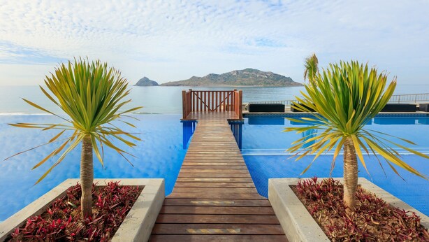 A wooden deck leading to a infinity pool with a view of the sea and a distant island.