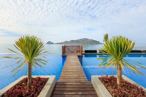 Eine Holzterrasse führt zu einem Infinity-Pool mit Blick auf das Meer und eine entfernte Insel.