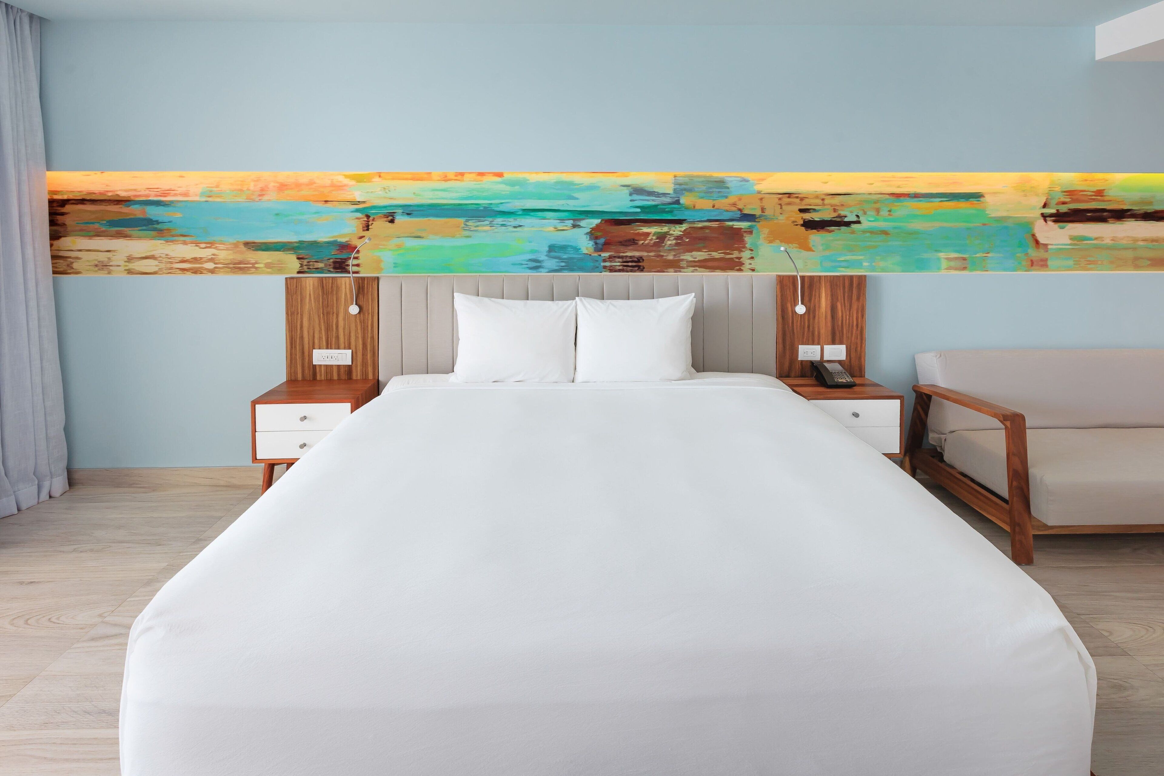 Une chambre d’hôtel moderne avec un grand lit, une tête de lit en bois, du linge de lit blanc, une table de chevet en bois avec un téléphone, et une peinture abstraite colorée accrochée au mur.