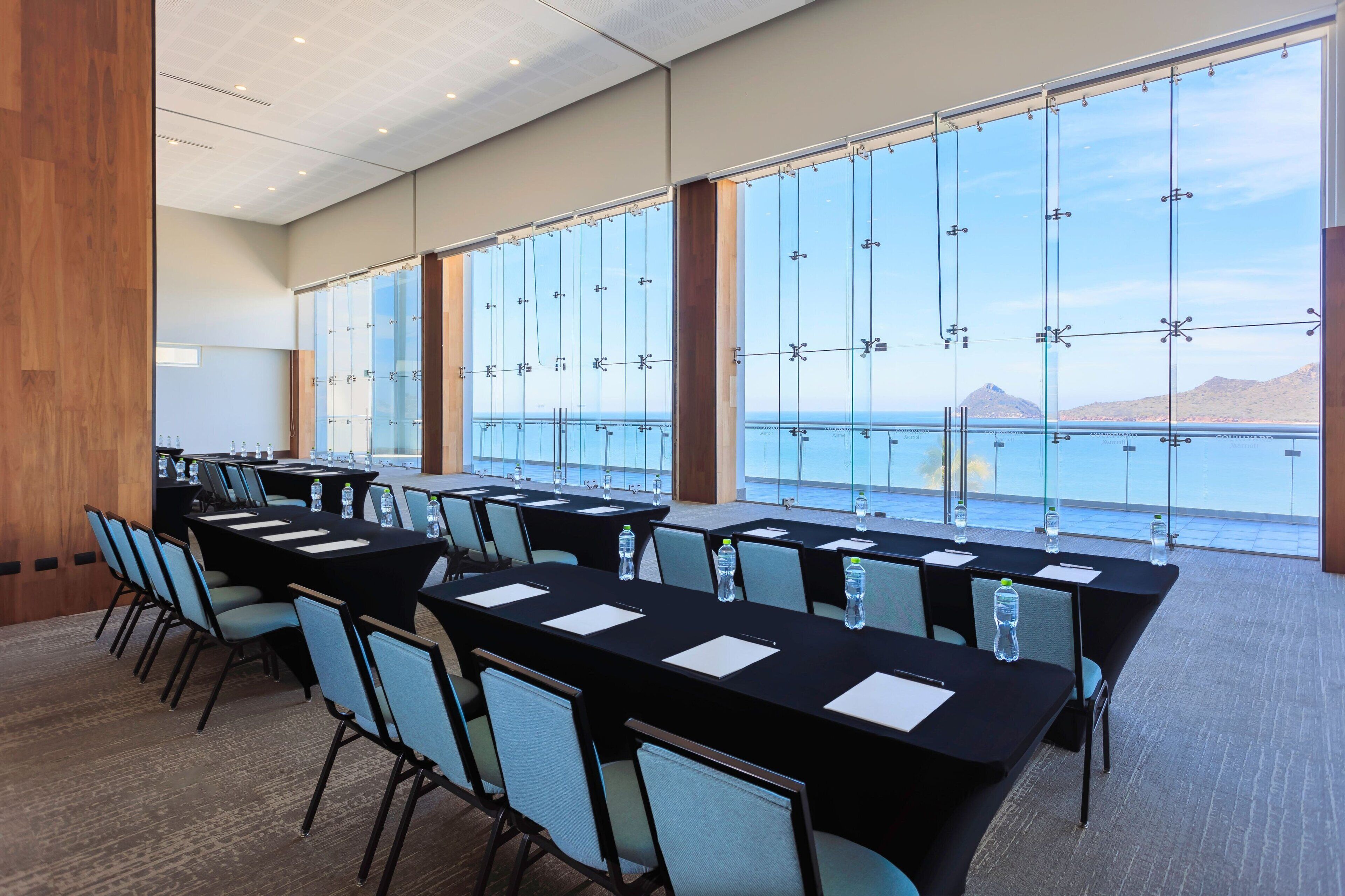 Une salle de conférence dotée de plusieurs tables et chaises, de grandes fenêtres offrant une vue sur la mer et les montagnes, et d’un panneau en bois sur le mur gauche.