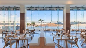 A restaurant with a long bench seating area, tables, and chairs, overlooking a scenic view of mountains and palm trees.