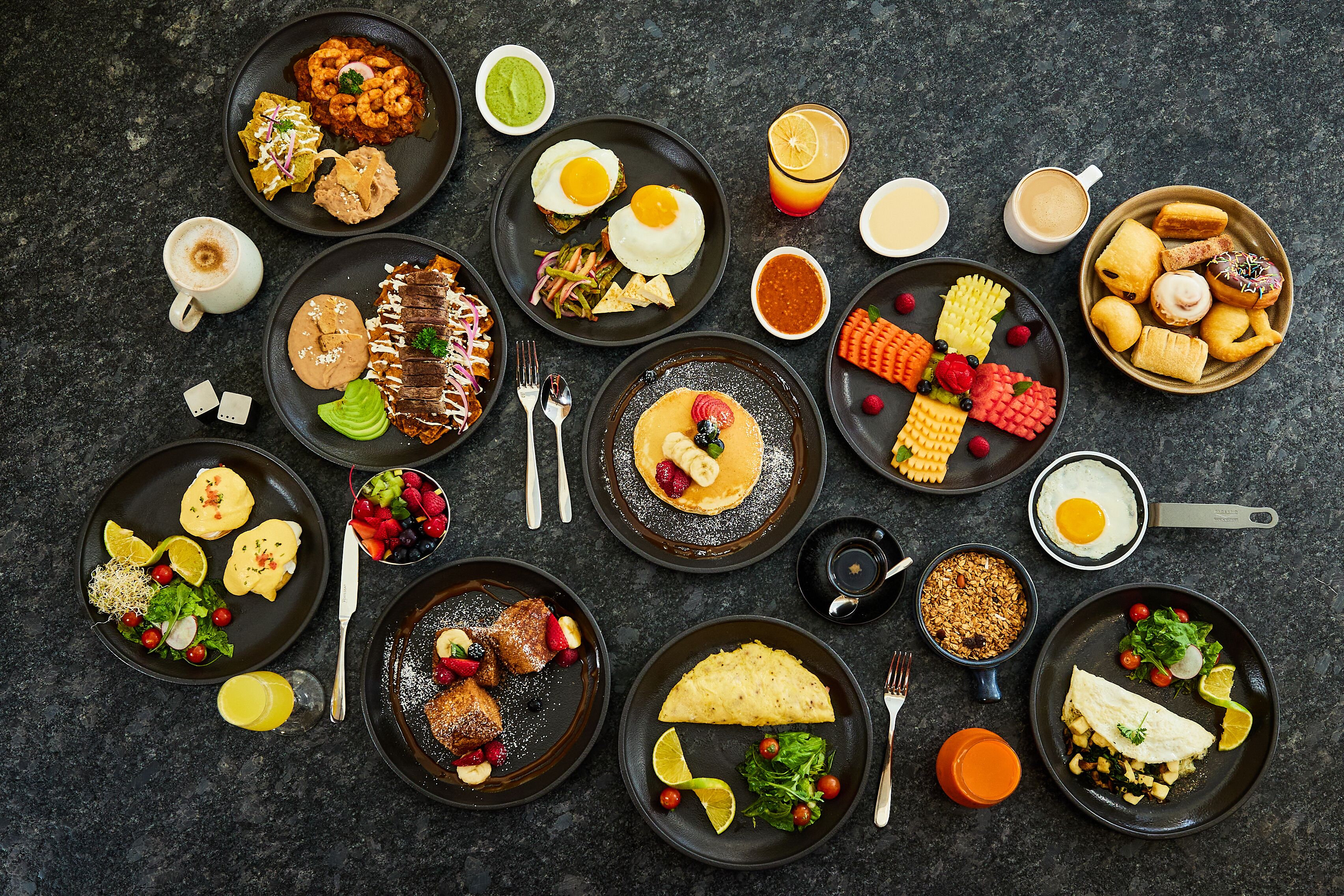 Une variété de plats pour le petit-déjeuner, présentés sur des assiettes et dans des bols, comprenant des œufs, des crêpes et des viennoiseries, le tout disposé sur une surface sombre.