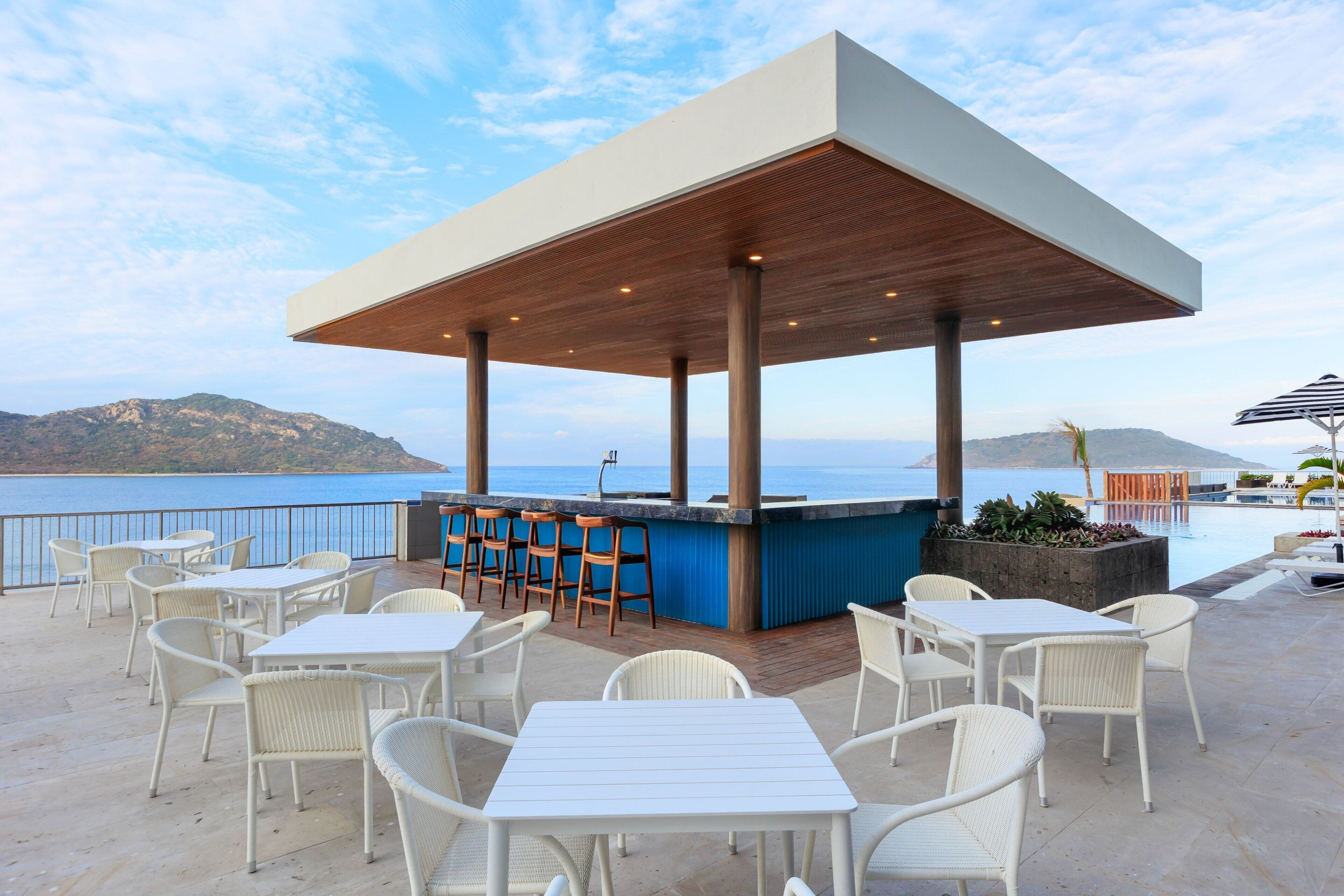 An outdoor terrace with white wicker furniture, a wooden bar, and a view of the sea.
