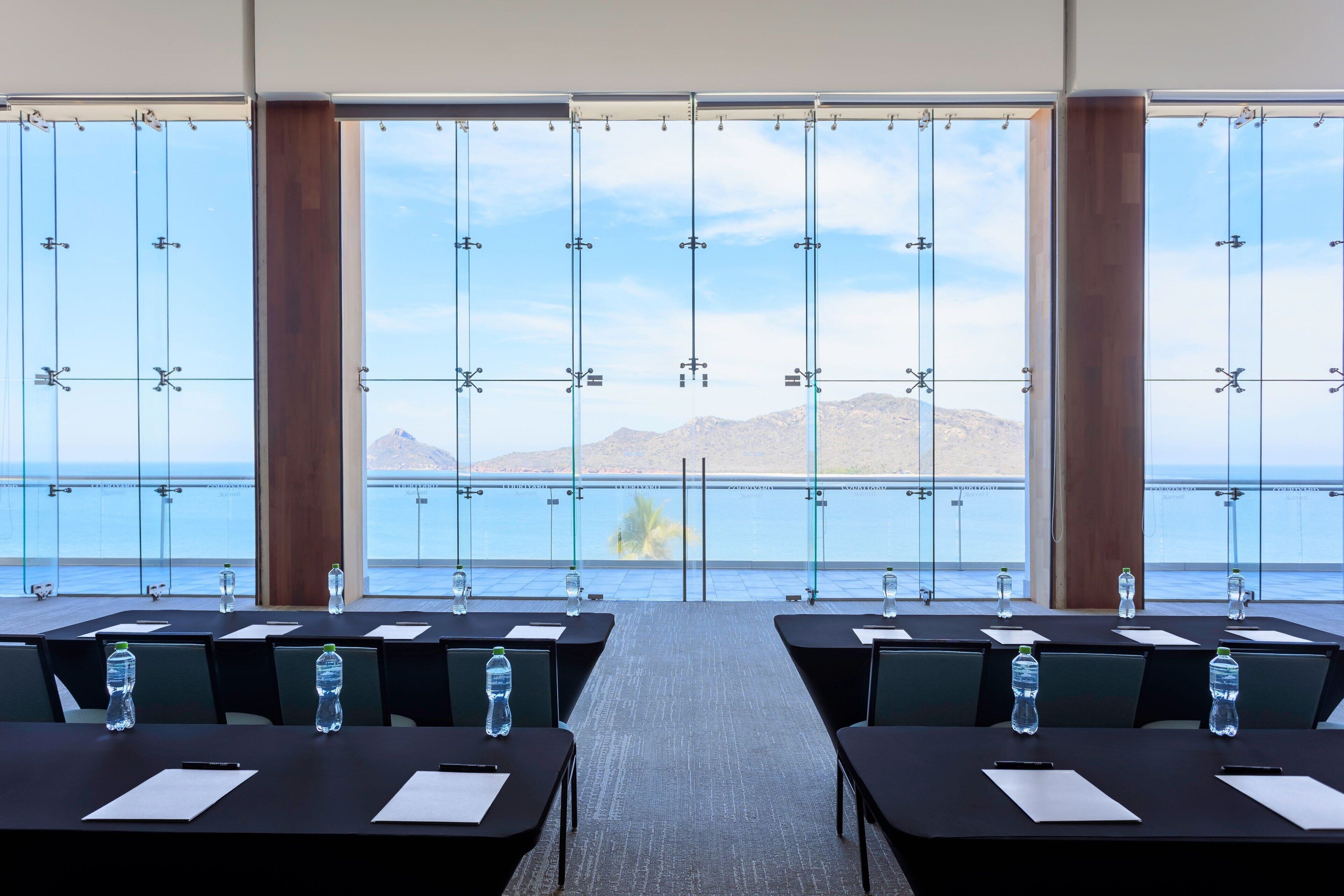 A conference room with tables, chairs, and water bottles. It has large windows with a view of the sea and mountains.