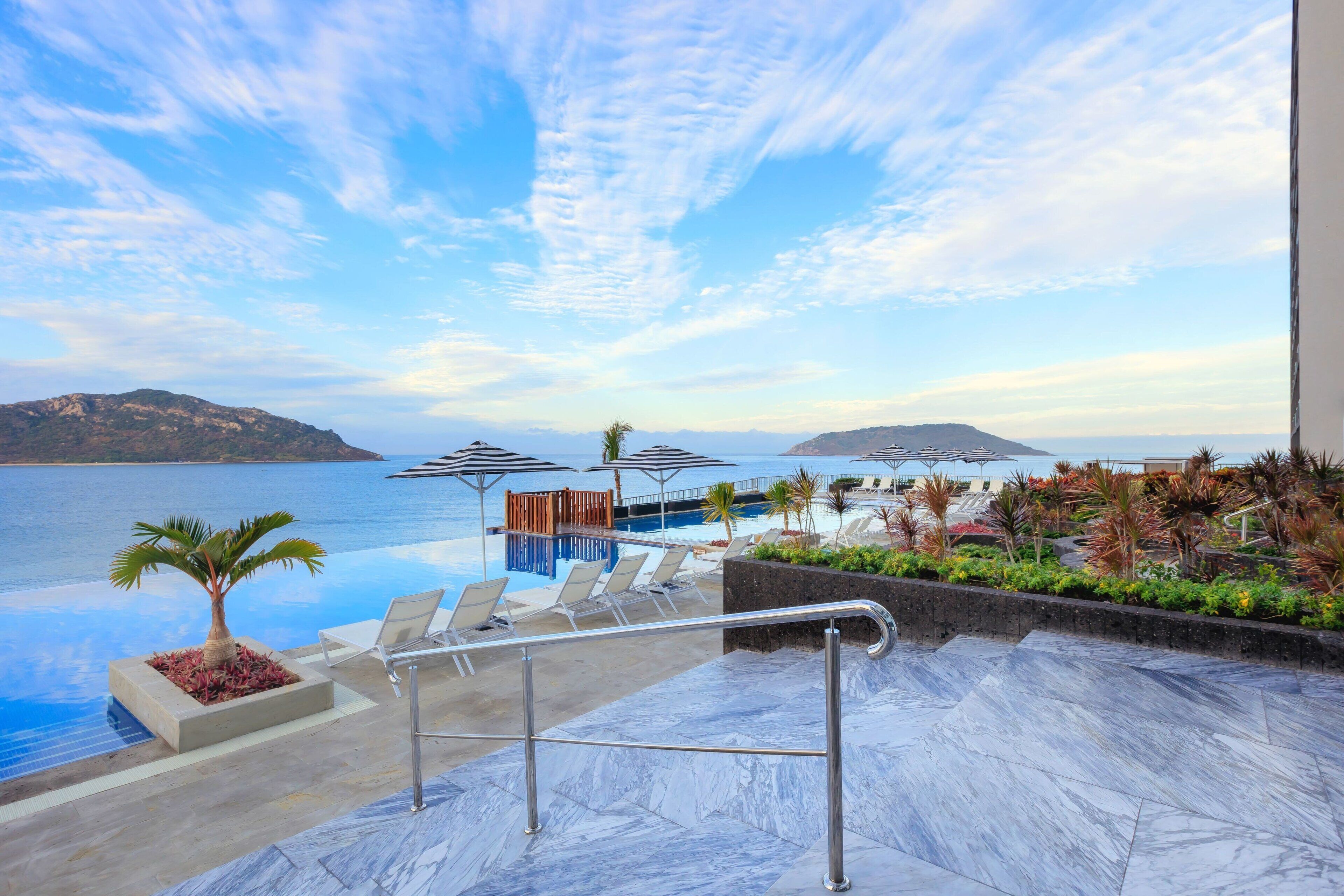 A pool area with lounge chairs, a palm tree, and a view of the sea.