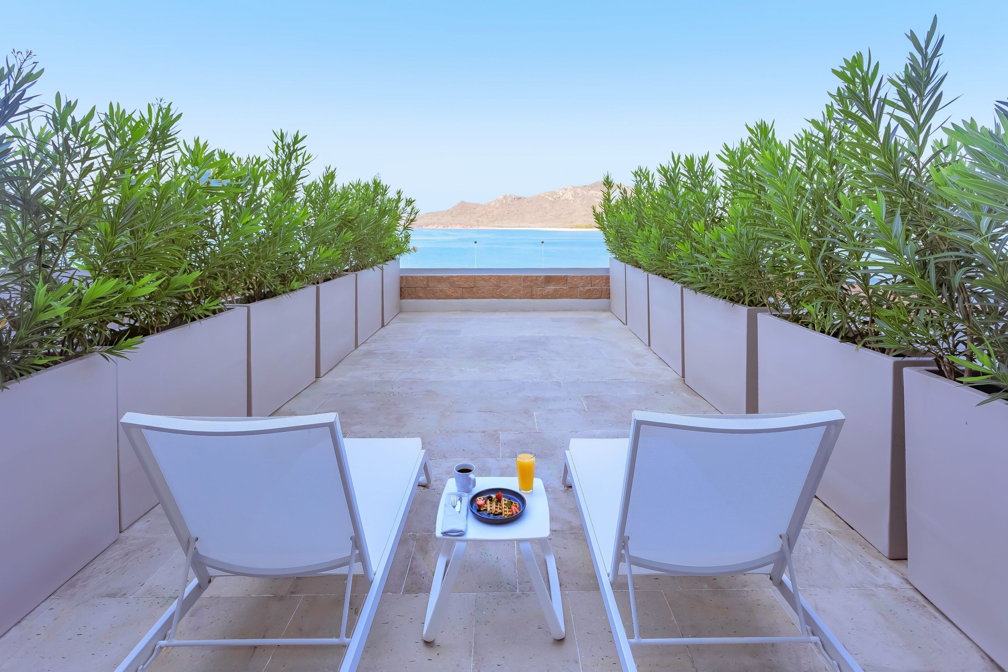 Un patio con dos sillas blancas y una mesa pequeña con un plato de comida, una taza y una botella. El patio tiene vista a un cuerpo de agua y está rodeado de follaje.