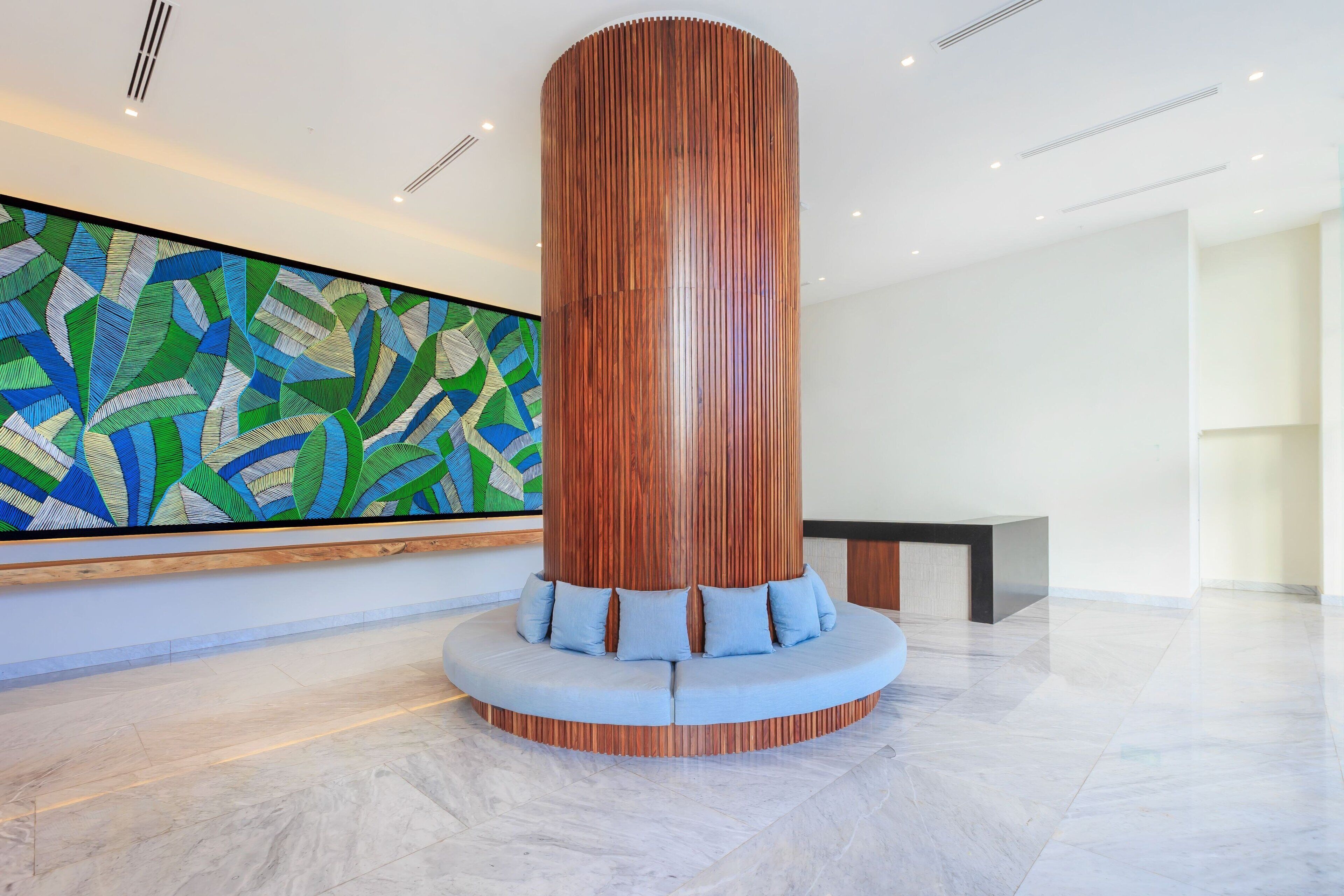 A modern lobby with a wooden cylindrical column, a seating area with blue cushions, and a colorful abstract mural on the wall.