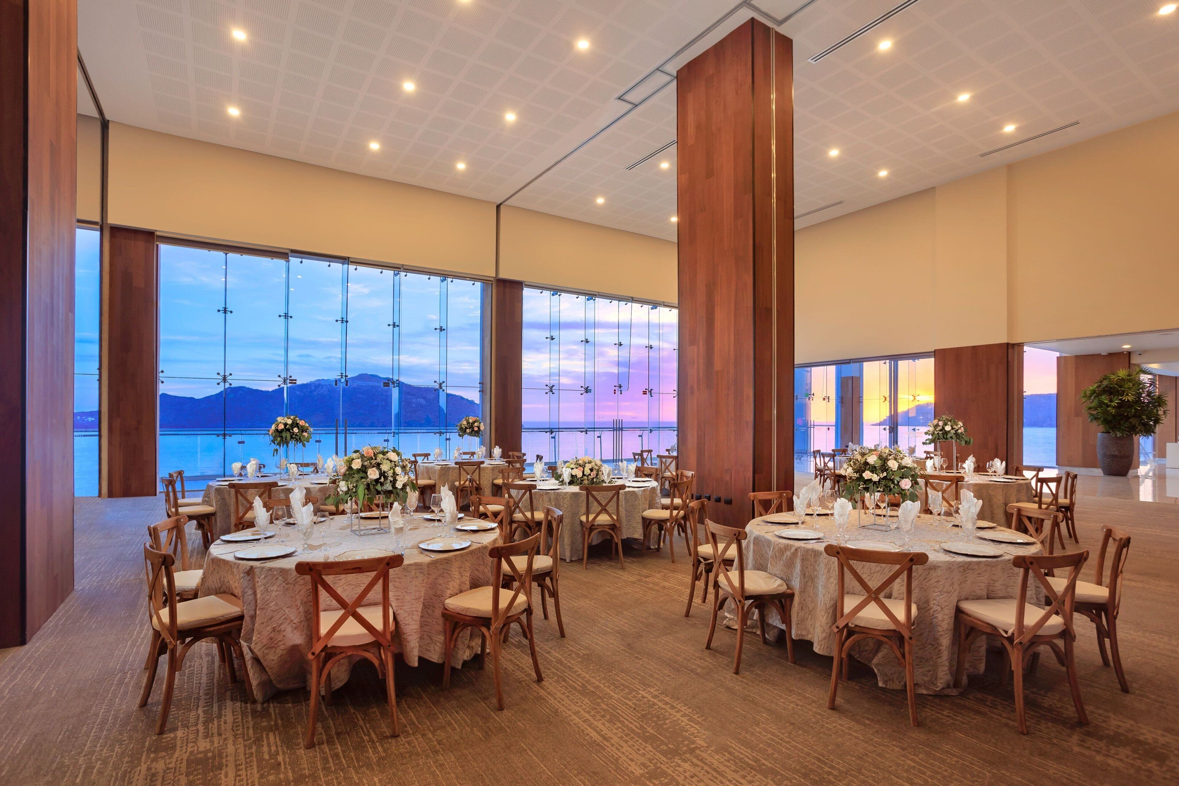 A spacious dining area with round tables set for a meal, large windows offering a view of the sea, and wooden paneling on the walls.