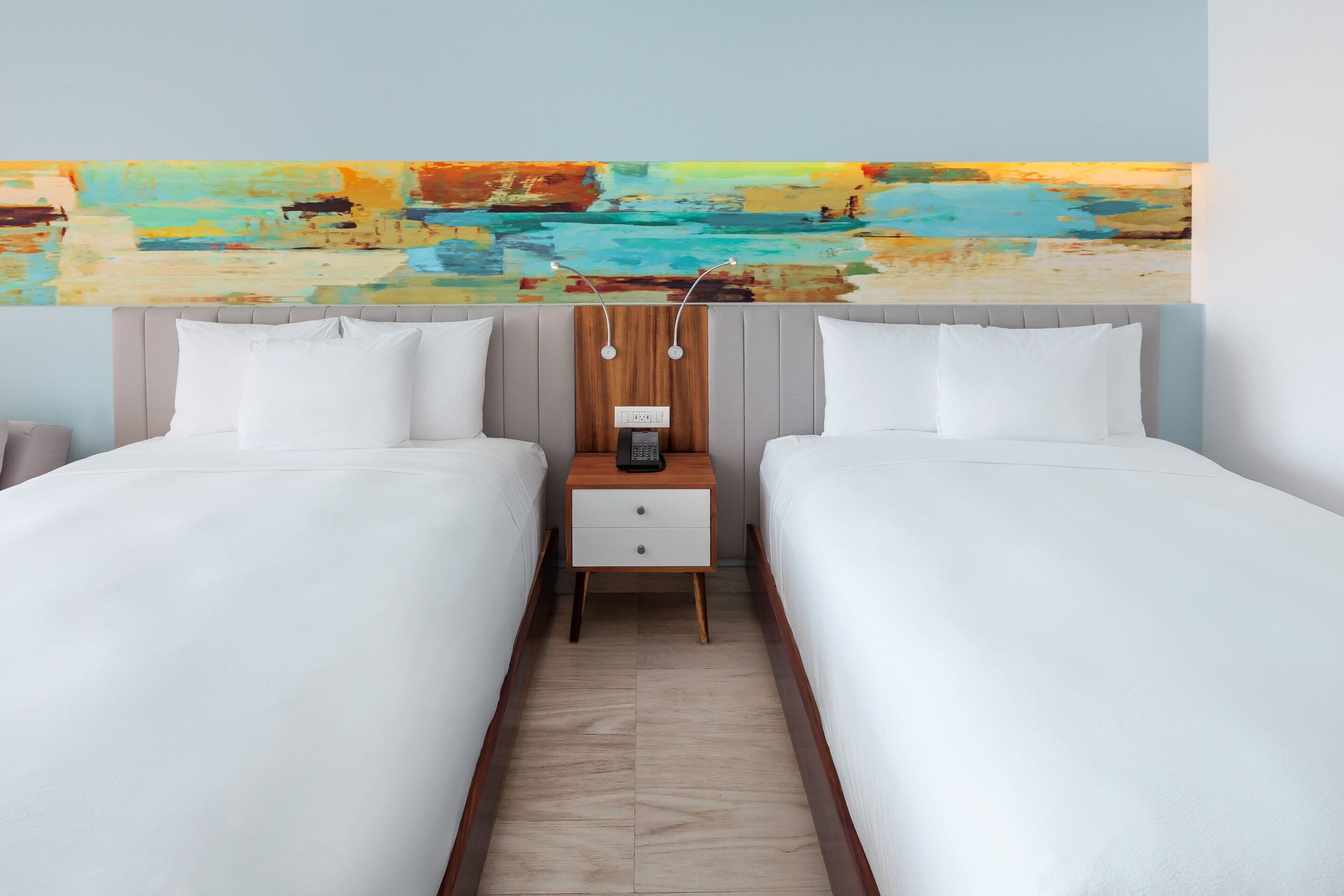 Habitación de hotel con dos camas, una mesita de noche de madera con un teléfono y un cuadro abstracto colorido sobre las camas.