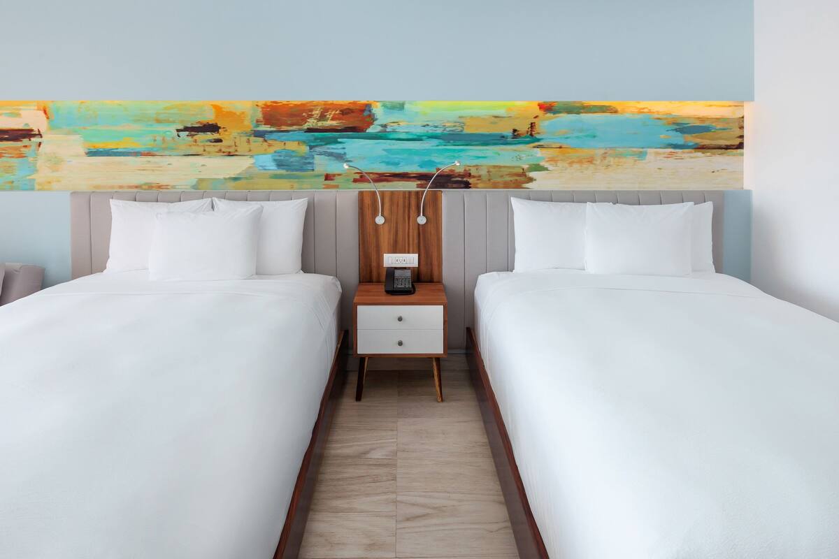 Habitación de hotel con dos camas, una mesita de noche de madera con un teléfono y un cuadro abstracto colorido sobre las camas.