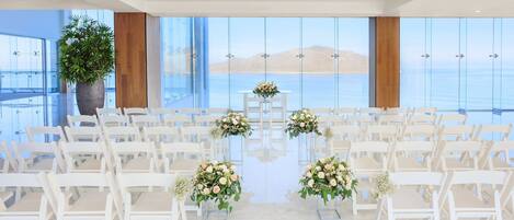 A wedding ceremony setup with white chairs and floral centerpieces, large windows with a view of the sea, and a potted plant.