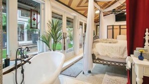 Une chambre avec une grande fenêtre, une baignoire blanche, un lit à baldaquin et une coiffeuse avec un miroir.