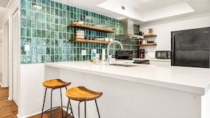 A modern kitchen with a white countertop, green tiled backsplash, wooden bar stools, and a black refrigerator. - 🏖️Garden View Kona Condo | A/C, Pools, Walk to Ocean (Kailua Kona)