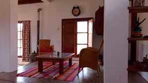 A room with a wooden table, two wicker chairs, a colorful rug, a wooden door, and a clock on the wall. - Casa Luciérnaga, ideal to enjoy the north of Argentina (humahuaca)