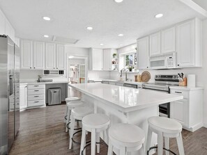 A modern kitchen with white cabinetry, a large island with bar stools, stainless steel appliances, and recessed ceiling lighting.