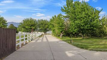 A paved pathway flanked by a white fence and trees, leading towards distant mountains.