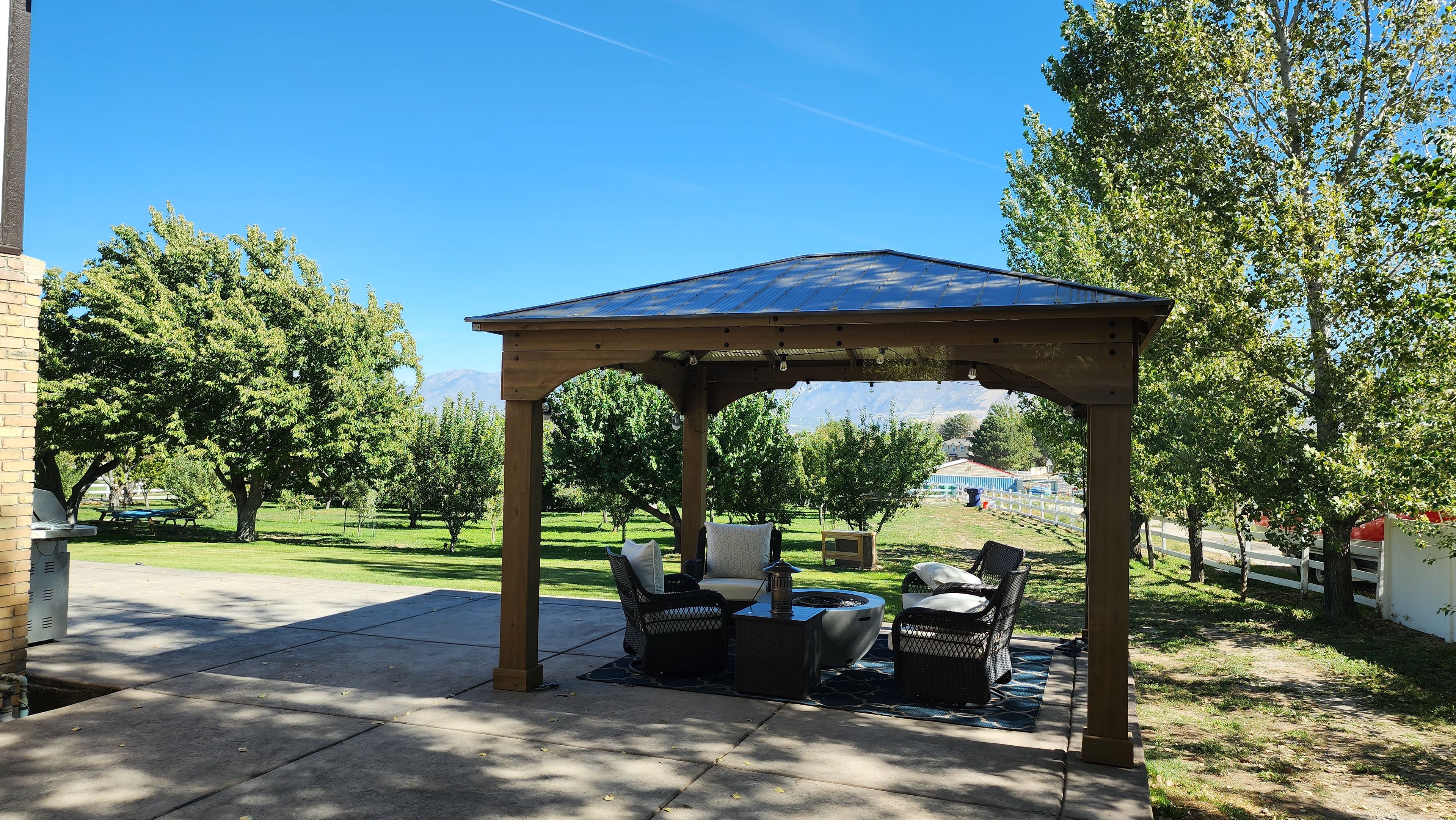 Un belvédère doté d’un toit, entouré de mobilier d’extérieur, et offrant une vue sur une zone verdoyante avec des arbres et un bâtiment situé au loin.