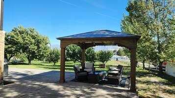 Un belvédère doté d’un toit, entouré de mobilier d’extérieur, et offrant une vue sur une zone verdoyante avec des arbres et un bâtiment situé au loin.