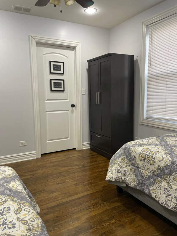 A bedroom with two beds, a dark wardrobe, a white door with framed pictures, and a window with blinds. - Woodlawn Oasis...10 Minutes to Dining & Downtown (Chicago)