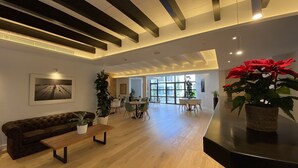 A spacious living area with a wooden floor, a dark brown sofa, a wooden bench, and a dining table with chairs. There is a large window providing natural light and a view of the outdoors. - Alaró Natura Rural Suites (Alaro)