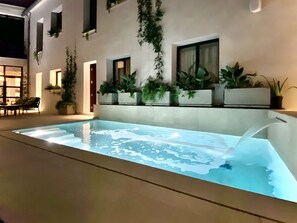 An indoor swimming pool with a clear water surface, surrounded by potted plants and a seating area with chairs.