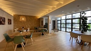 A spacious dining area with wooden tables and chairs, a stone accent wall, and large windows offering a view of the outside. - Alaró Natura Rural Suites (Alaro)