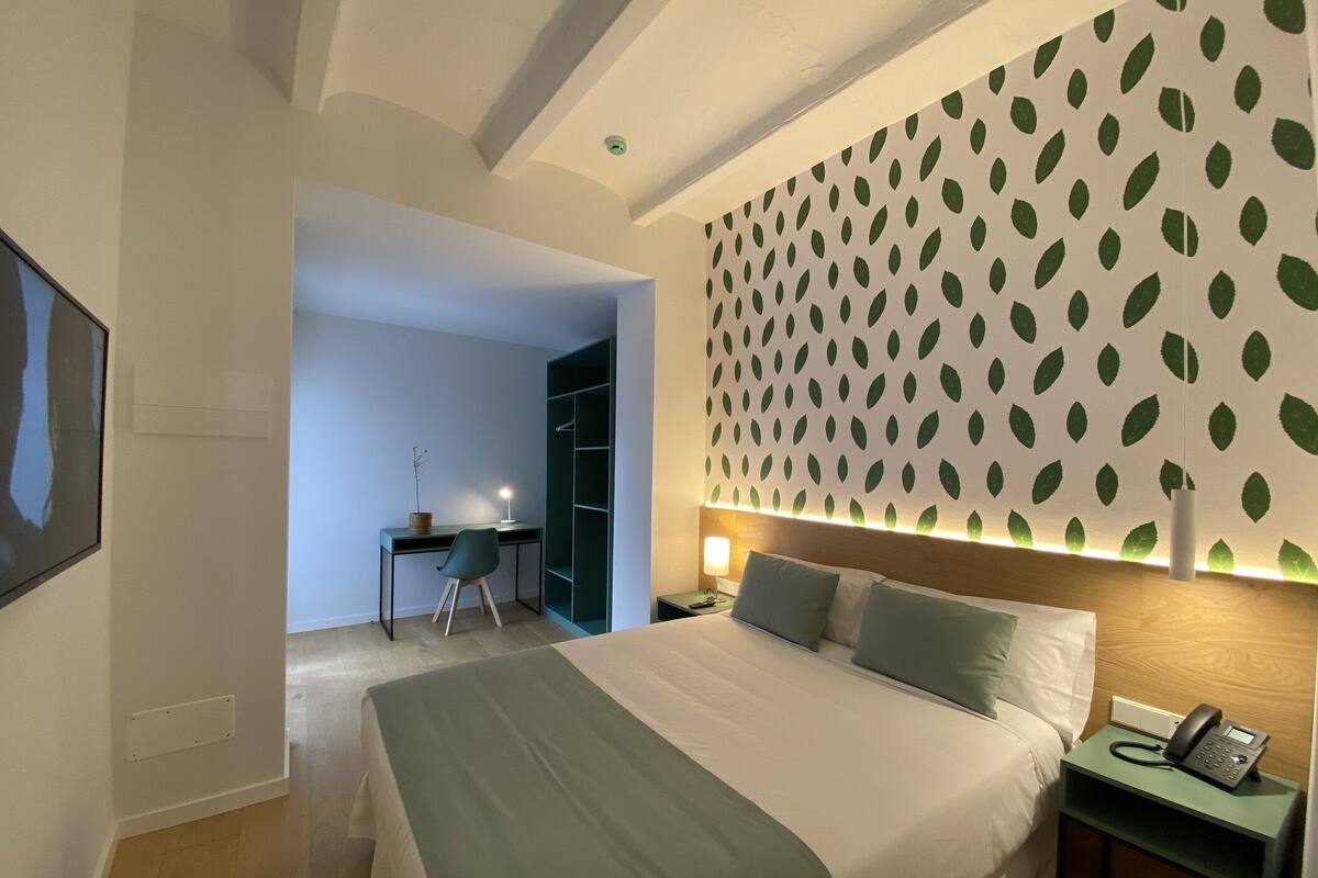 Quarto de hotel com cama, escrivaninha e papel de parede com padrão de folhas verdes.