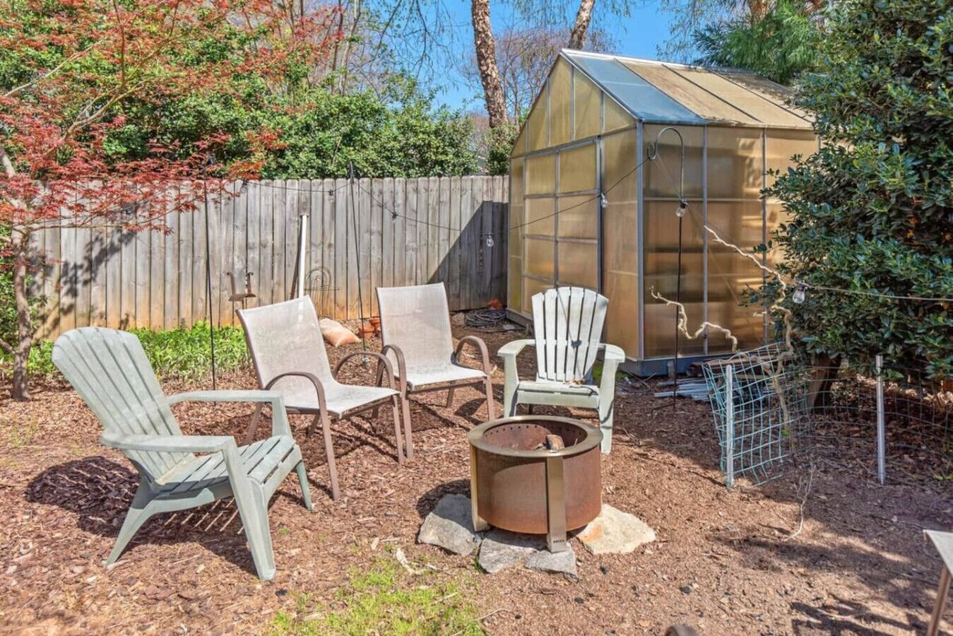 Un patio trasero con valla de madera, un invernadero de vidrio y cuatro sillas Adirondack alrededor de un brasero.