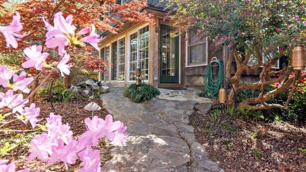 A stone pathway leading to a wooden house with large windows and a garden with pink flowers and trees.