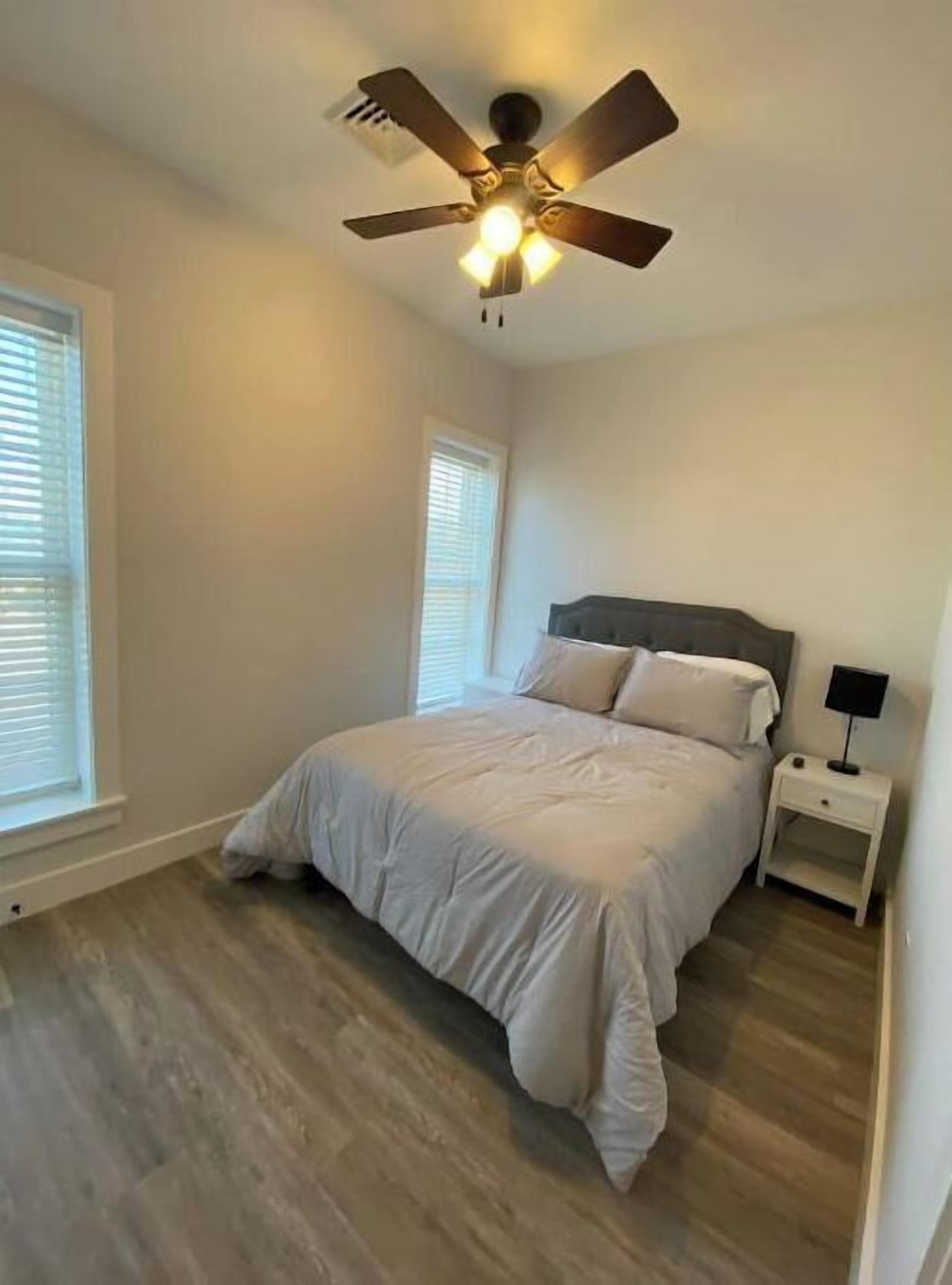 A bedroom with a bed, a nightstand, a ceiling fan, and two windows with blinds.