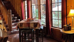 A wooden interior with a dining table, chairs, a staircase, and large windows with curtains.
