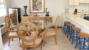 A dining area with a glass-top table surrounded by wooden chairs with cushioned seats, a kitchen area with a white countertop and blue bar stools, and a framed painting on the wall. - Beach Front (Rincón)