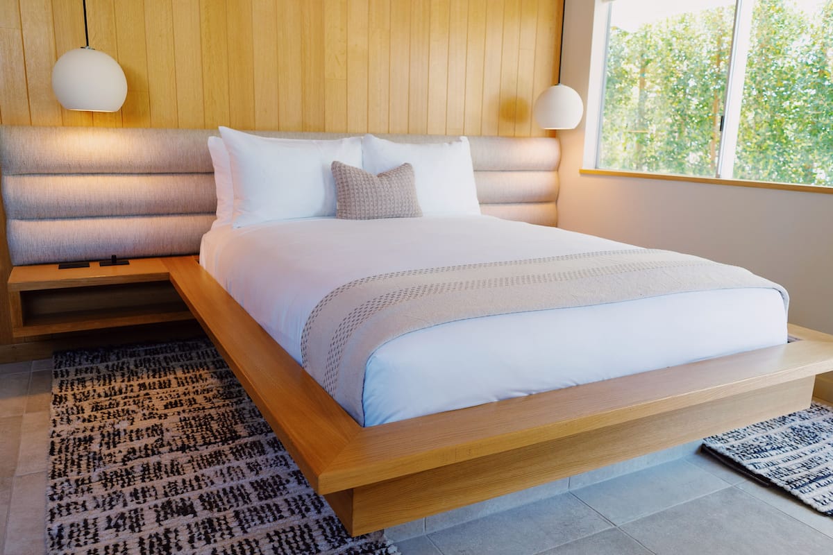 Un dormitorio moderno con cabecera de madera, una cama grande con ropa de cama blanca y vistas a árboles a través de una ventana.