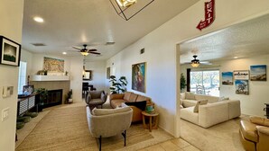 A spacious living room with a fireplace, sofas, and a ceiling fan. - Epic Views, Huge House, Acres of J-Trees to explore + Hot Tub, Pool, AC & Golf! (Joshua Tree)