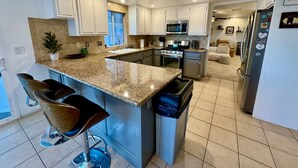 A modern kitchen with granite countertops, stainless steel appliances, and a central island with bar stools. - Epic Views, Huge House, Acres of J-Trees to explore + Hot Tub, Pool, AC & Golf! (Joshua Tree)