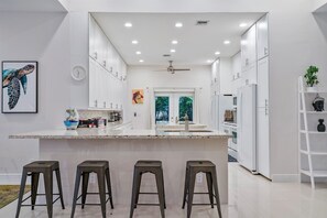 Une cuisine moderne avec des meubles blancs, un îlot central doté de tabourets, un ventilateur de plafond et une fenêtre donnant sur un espace verdoyant.