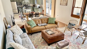 Ein geräumiges Wohnzimmer mit einer braunen Ledersofa, einem grünen Sofa und einem Couchtisch aus Holz. Es gibt einen Essbereich mit Tisch und Stühlen sowie eine Küche im Hintergrund.