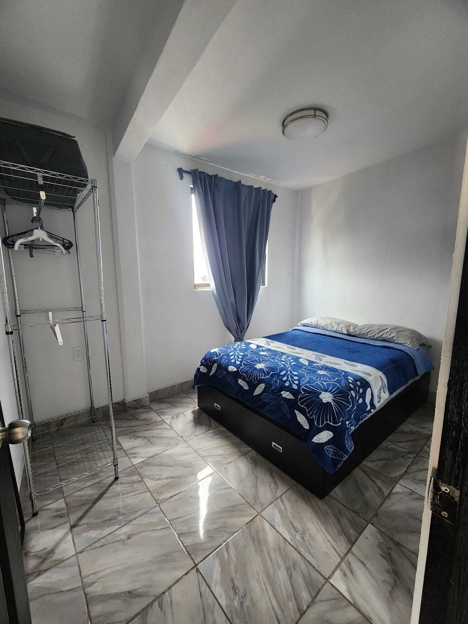 A bedroom with a bed, a window with curtains, a metal drying rack, and a tiled floor.