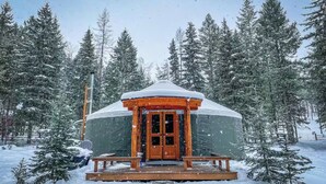 Une yourte en bois, dont le toit est recouvert de neige, entourée d’arbres chargés de neige.