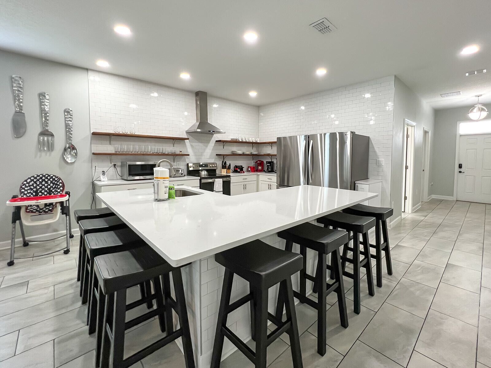 Una cocina moderna con una isla grande, taburetes negros, electrodomésticos de acero inoxidable y azulejos blancos tipo metro.
