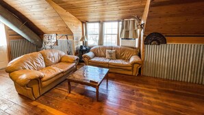 A cozy loft-style room with wooden floors, a leather sofa, a coffee table, and a large window with curtains. - Quiet Crystal Creek Ranch Bunkhouse with large private pond (Brackettville)