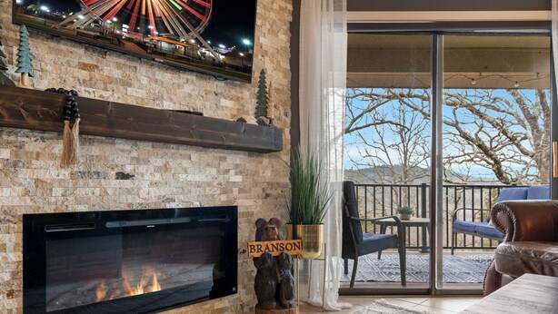 A fireplace with a bear decoration, a Ferris wheel in the background, and a balcony with chairs. - Woodsy Wonder - Pools, Hot tub, Gated & Golf CENTRAL LOCATION (Branson)