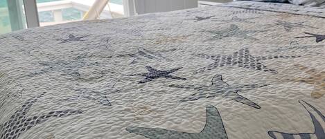 A bedroom with a bed featuring a starfish-patterned quilt, a view of the ocean through a window, and a white lamp on a bedside table.