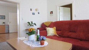 Una sala de estar con un sofá rojo, una mesa de centro de madera con una botella y flores, y una puerta blanca al fondo.