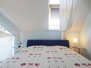Un dormitorio con cama, ventana con cortinas, lámpara y una mesita.