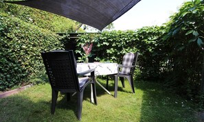 An outdoor patio with a table and two chairs, shaded by a large umbrella, surrounded by lush greenery. - Gartenblick - Willkommen in der Ferienwohnung Gartenblick in Kröpelin (Kröpelin)
