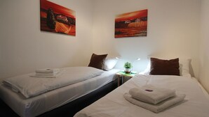 Een hotelkamer met twee bedden, elk voorzien van witte lakens en kussens, een klein nachtkastje met een plant, en twee schilderijen aan de muur.