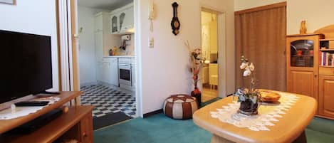 A living room with a television, a wooden coffee table with a lace doily, a patterned ottoman, and a kitchen in the background.