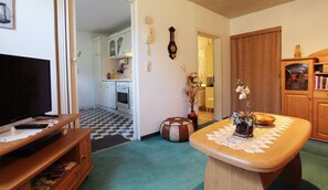 A living room with a television, a wooden coffee table with a lace doily, a patterned ottoman, and a kitchen in the background. - Gartenblick - Willkommen in der Ferienwohnung Gartenblick in Kröpelin (Kröpelin)